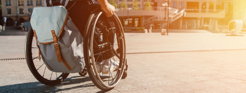 cropped panoramic view of man using wheelchair with bag on street with sunlight